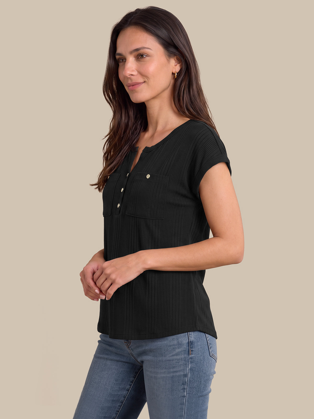 A woman wearing a black short-sleeve ribbed henley top with gold buttons and blue jeans, standing against a solid beige background.