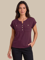 A woman modeling a short-sleeve ribbed henley top in deep plum purple with gold-tone buttons, paired with black pants, against a solid beige background.