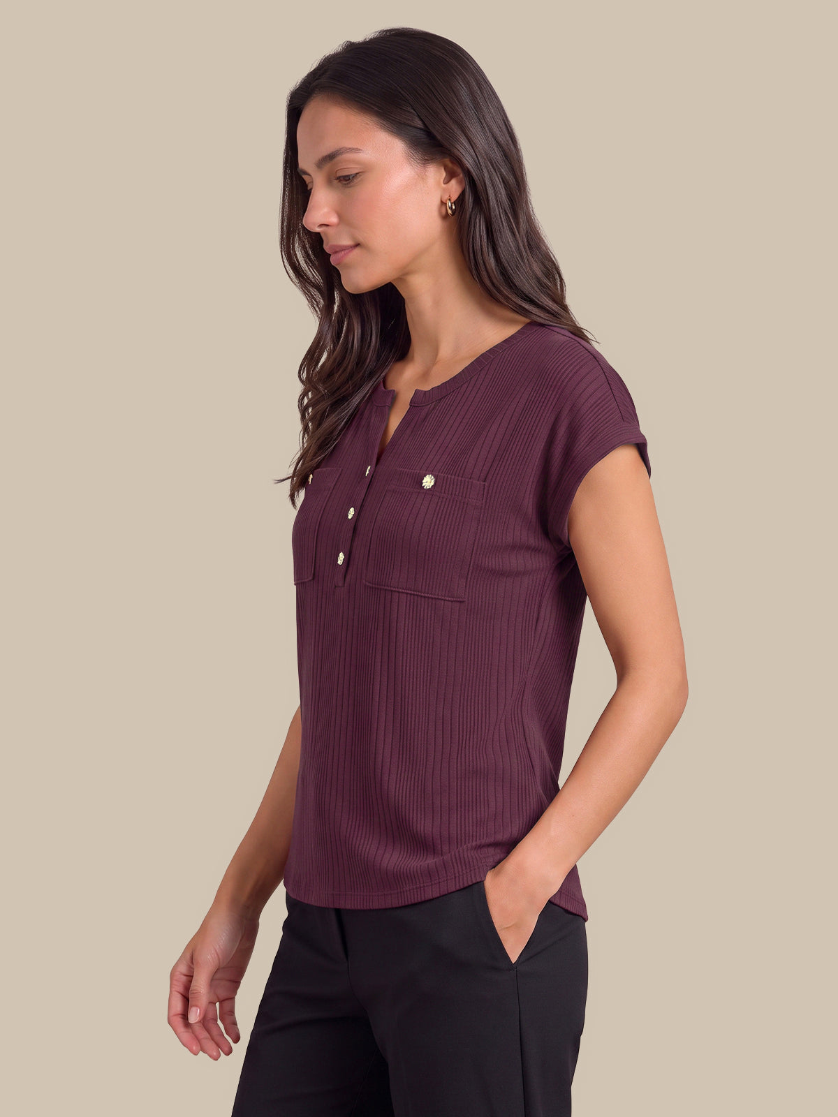 A woman wearing a short-sleeve ribbed henley top in deep plum purple with gold-tone buttons, paired with black pants, set against a solid beige background.