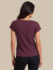 A woman shown from the back wearing a short-sleeve ribbed knit top in a deep plum purple color, paired with black pants, against a solid beige background.