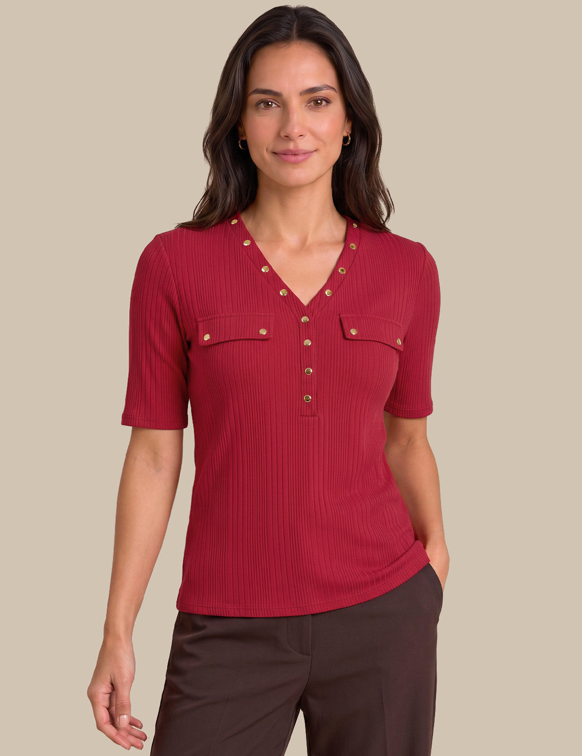 A woman modeling a short-sleeve ribbed knit top in deep red with gold-tone decorative buttons and chest pockets, paired with dark brown pants, against a solid beige background.