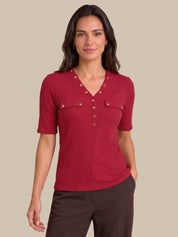A woman modeling a short-sleeve ribbed knit top in deep red with gold-tone decorative buttons and chest pockets, paired with dark brown pants, against a solid beige background.
