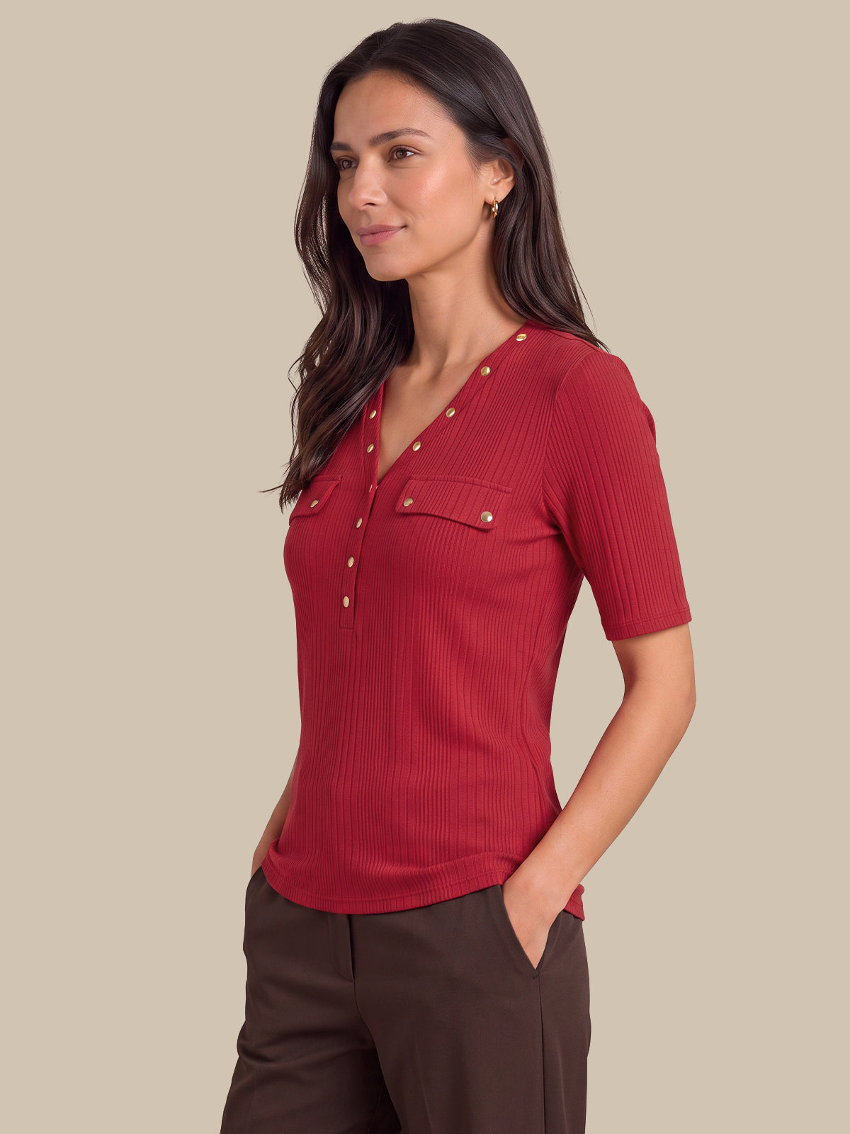 A woman wearing a short-sleeve ribbed knit top in deep red with gold-tone decorative buttons, paired with dark brown trousers, set against a solid beige background.