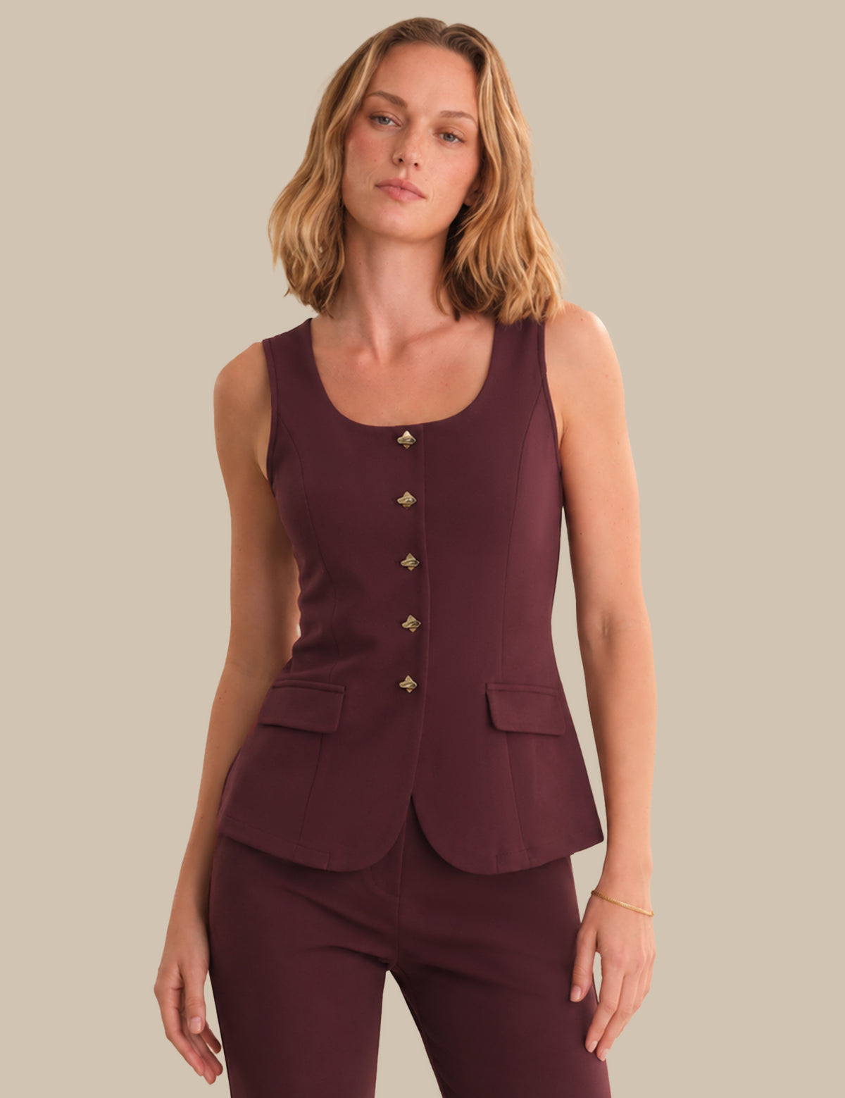 Woman wearing a maroon sleeveless top with buttons and matching pants against a beige background