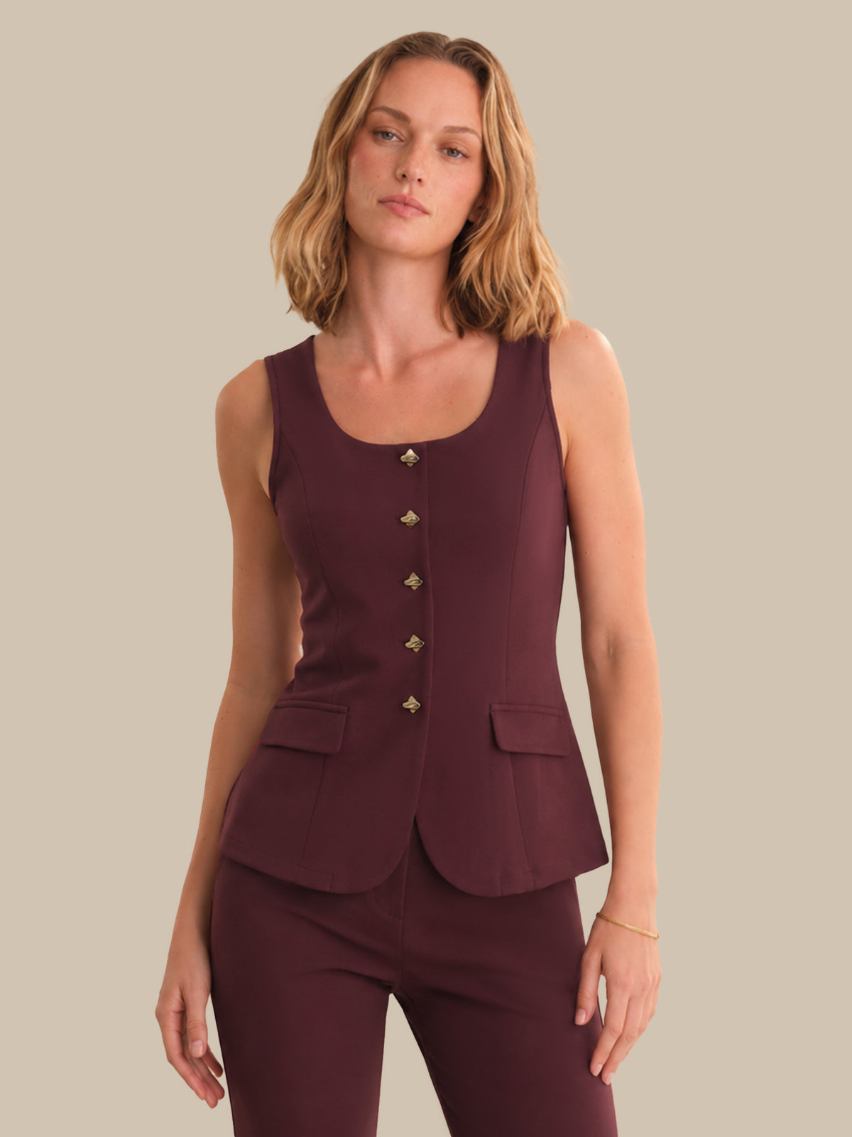Woman wearing a maroon sleeveless top with buttons and matching pants against a beige background