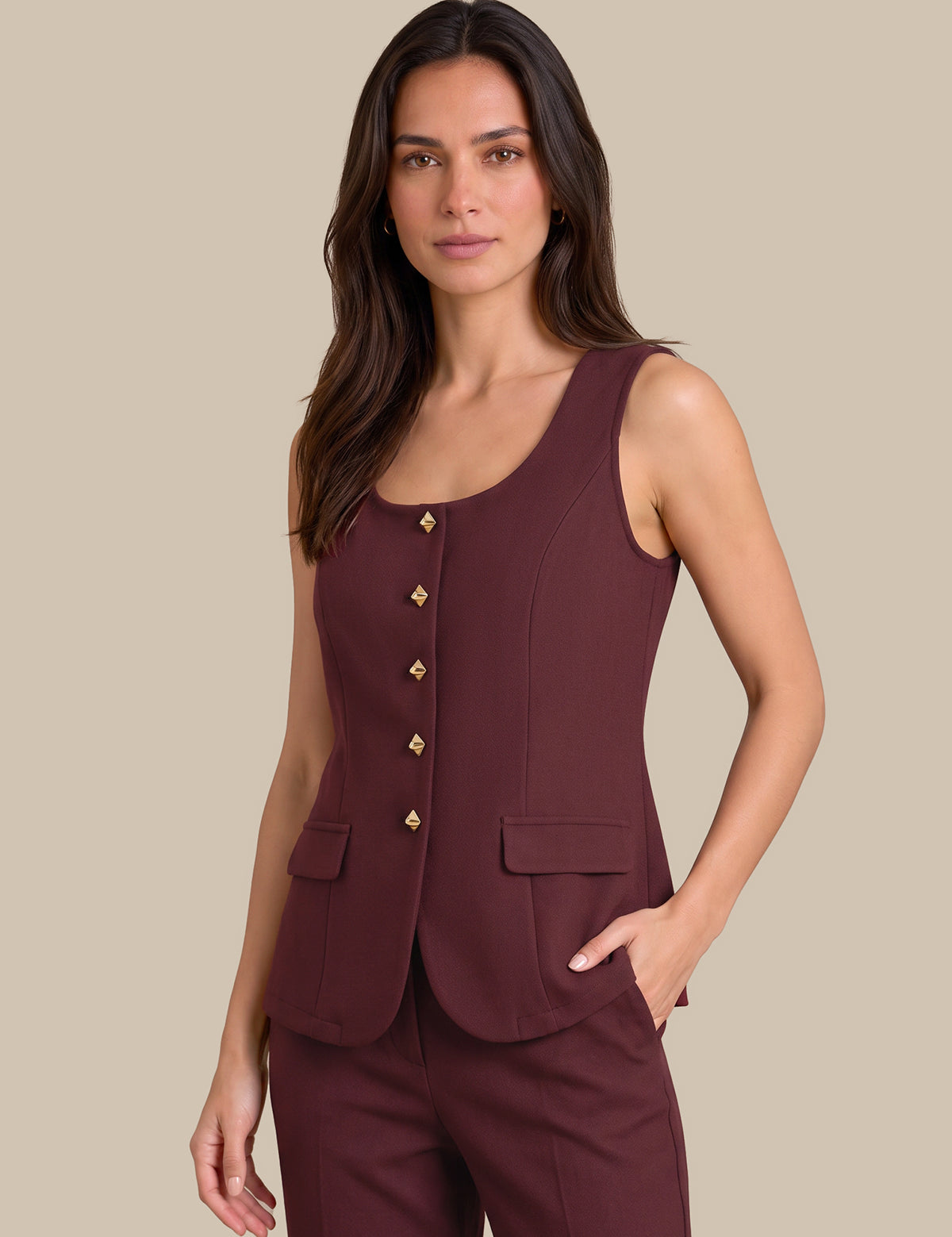 A woman modeling a sleeveless scoop-neck vest in deep burgundy with gold-tone decorative buttons down the front, paired with matching trousers, against a solid beige background.