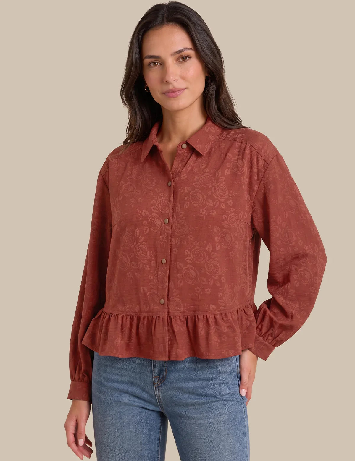 A woman modeling a rust-colored, long-sleeve button-up blouse with a subtle damask pattern and a ruffled hem, paired with blue jeans against a solid beige background.