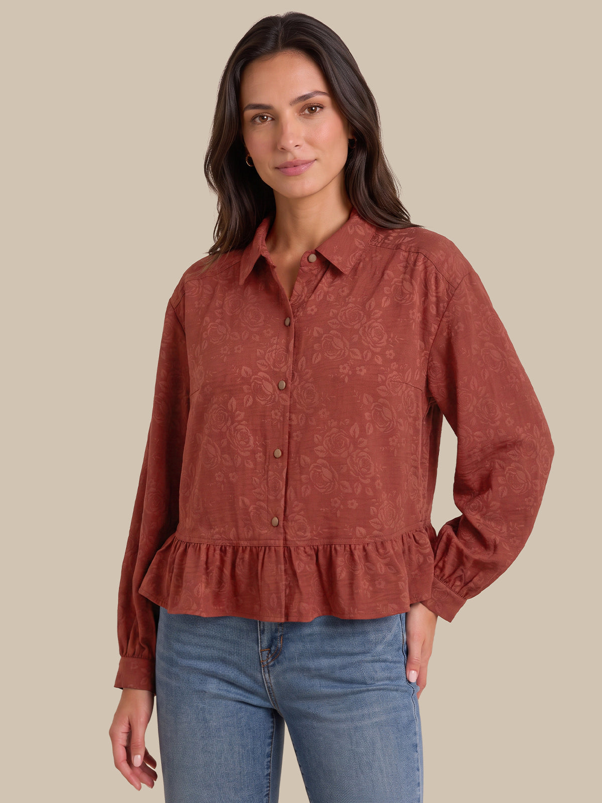 A woman modeling a rust-colored, long-sleeve button-up blouse with a subtle damask pattern and a ruffled hem, paired with blue jeans against a solid beige background.