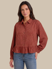 A woman modeling a rust-colored, long-sleeve button-up blouse with a subtle damask pattern and a ruffled hem, paired with blue jeans against a solid beige background.