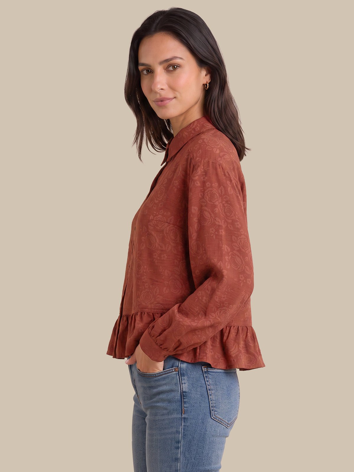 A woman wearing a rust-colored, long-sleeve textured blouse with a ruffled hem, paired with blue jeans, shown in a side-profile view against a solid beige background.
