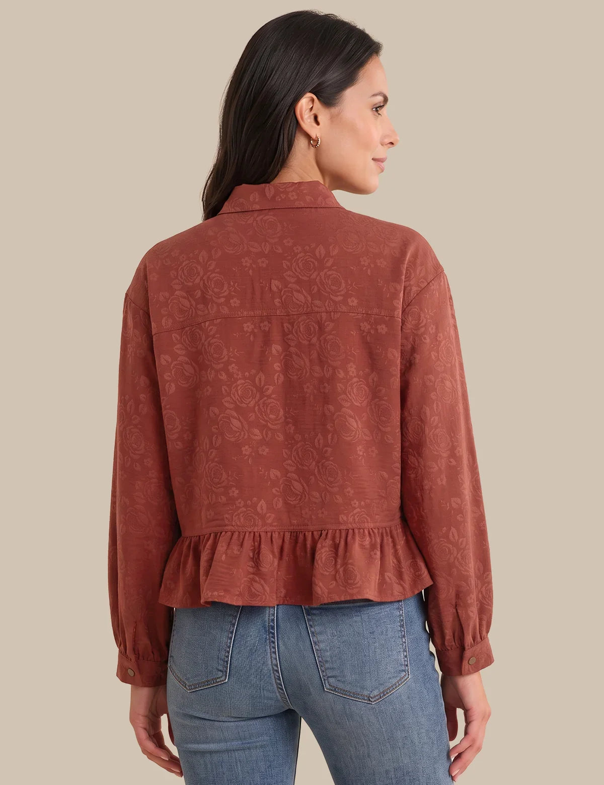 A woman shown from the back wearing a rust-colored long-sleeve blouse with a subtle tonal floral jacquard pattern and a ruffled hem, paired with blue jeans, against a solid beige background.