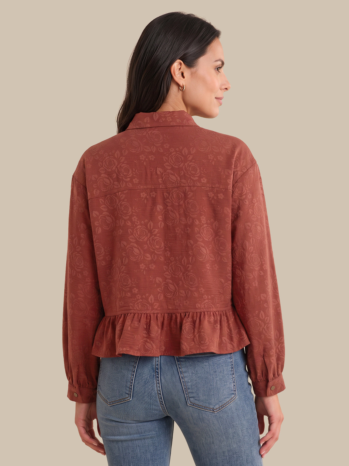 A woman shown from the back wearing a rust-colored long-sleeve blouse with a subtle tonal floral jacquard pattern and a ruffled hem, paired with blue jeans, against a solid beige background.