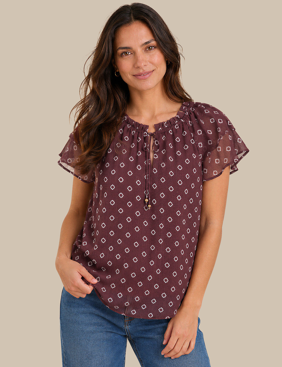 A woman wearing a short-sleeve blouse with a purple and white geometric pattern, paired with blue jeans, against a solid beige background.