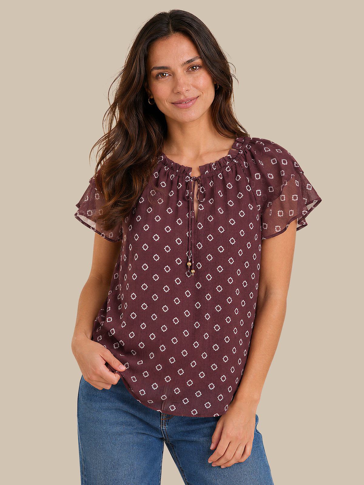 A woman wearing a short-sleeve blouse with a purple and white geometric pattern, paired with blue jeans, against a solid beige background.