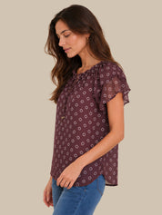 A woman wearing a short-sleeve, gathered blouse with a small white geometric pattern on a dark plum purple background, paired with blue jeans, shown against a solid beige background.