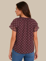 A woman shown from the back wearing a short-sleeve ruffled blouse in a deep plum purple with a small white geometric pattern, paired with blue jeans, against a solid beige background.