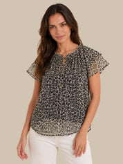 A woman modeling a sheer short-sleeve blouse with a black and gray leopard print, featuring a keyhole neckline with ruffle trim and white pants, set against a solid beige background.