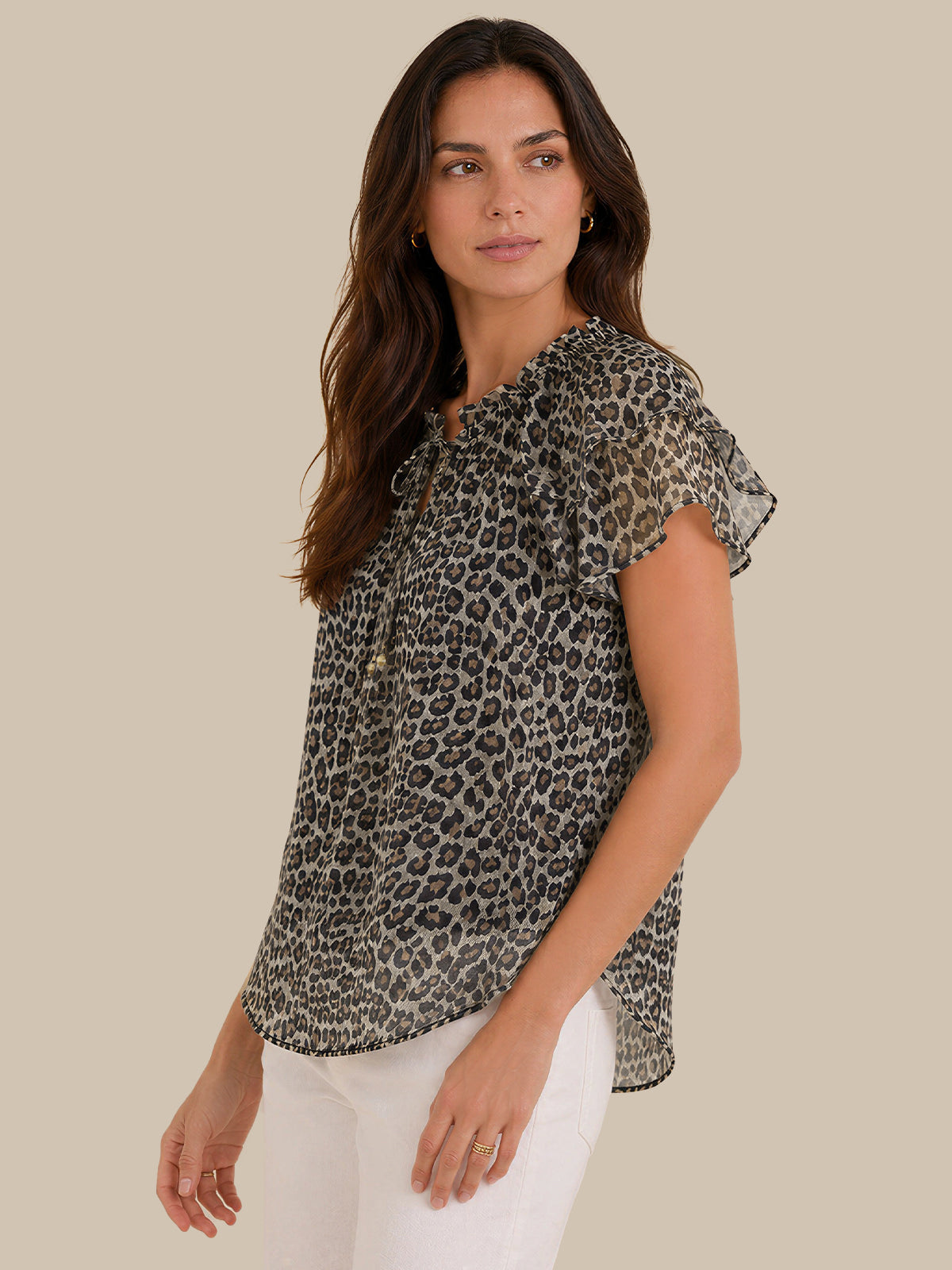 A woman modeling a short-sleeve leopard print blouse with a ruffled neckline and button-front detail, paired with white pants, against a solid beige background.