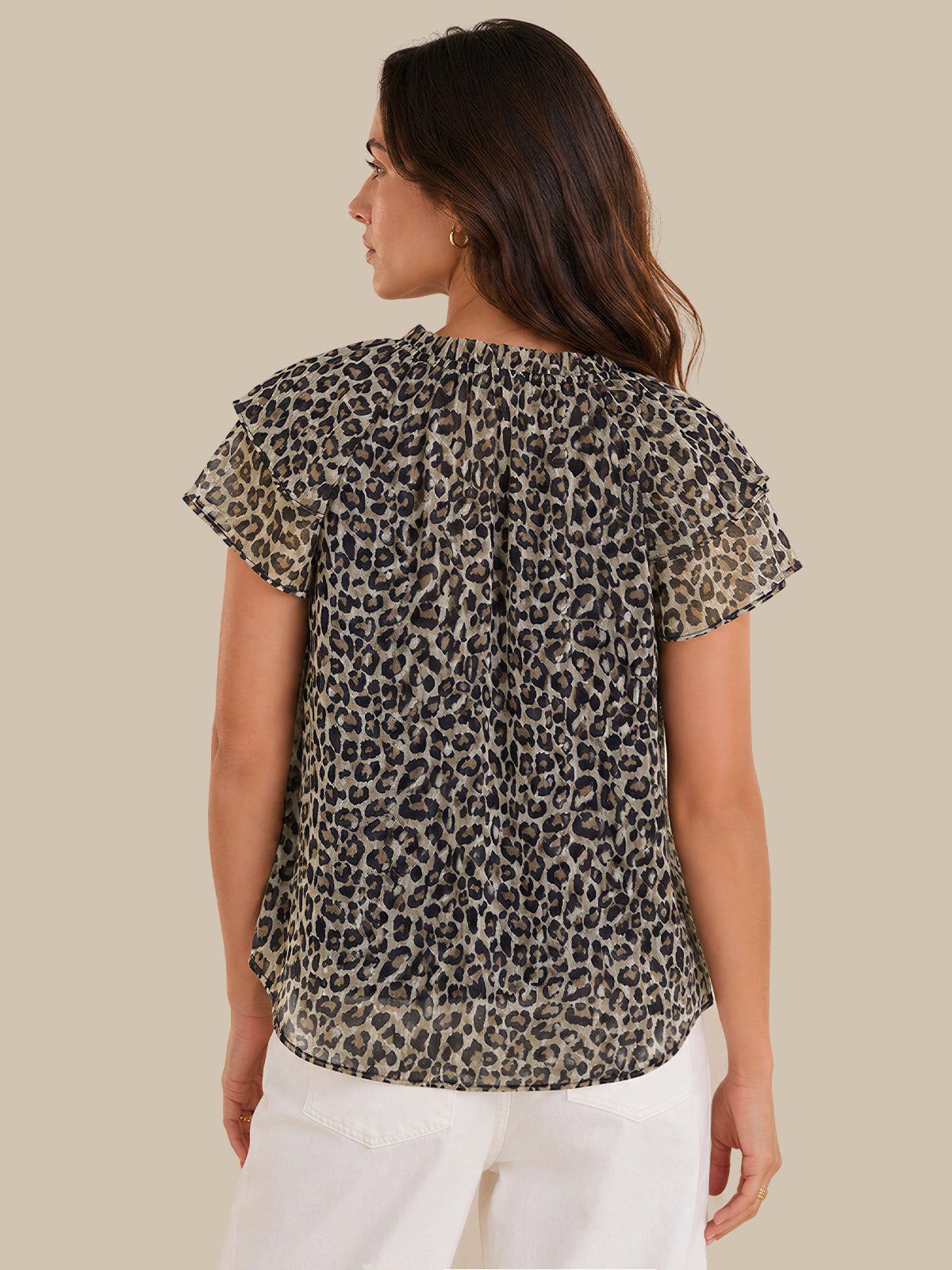 A woman shown from the back wearing a short-sleeve leopard print blouse with a gathered back and white pants, against a solid beige background.