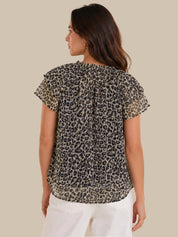 A woman shown from the back wearing a short-sleeve leopard print blouse with a gathered back and white pants, against a solid beige background.
