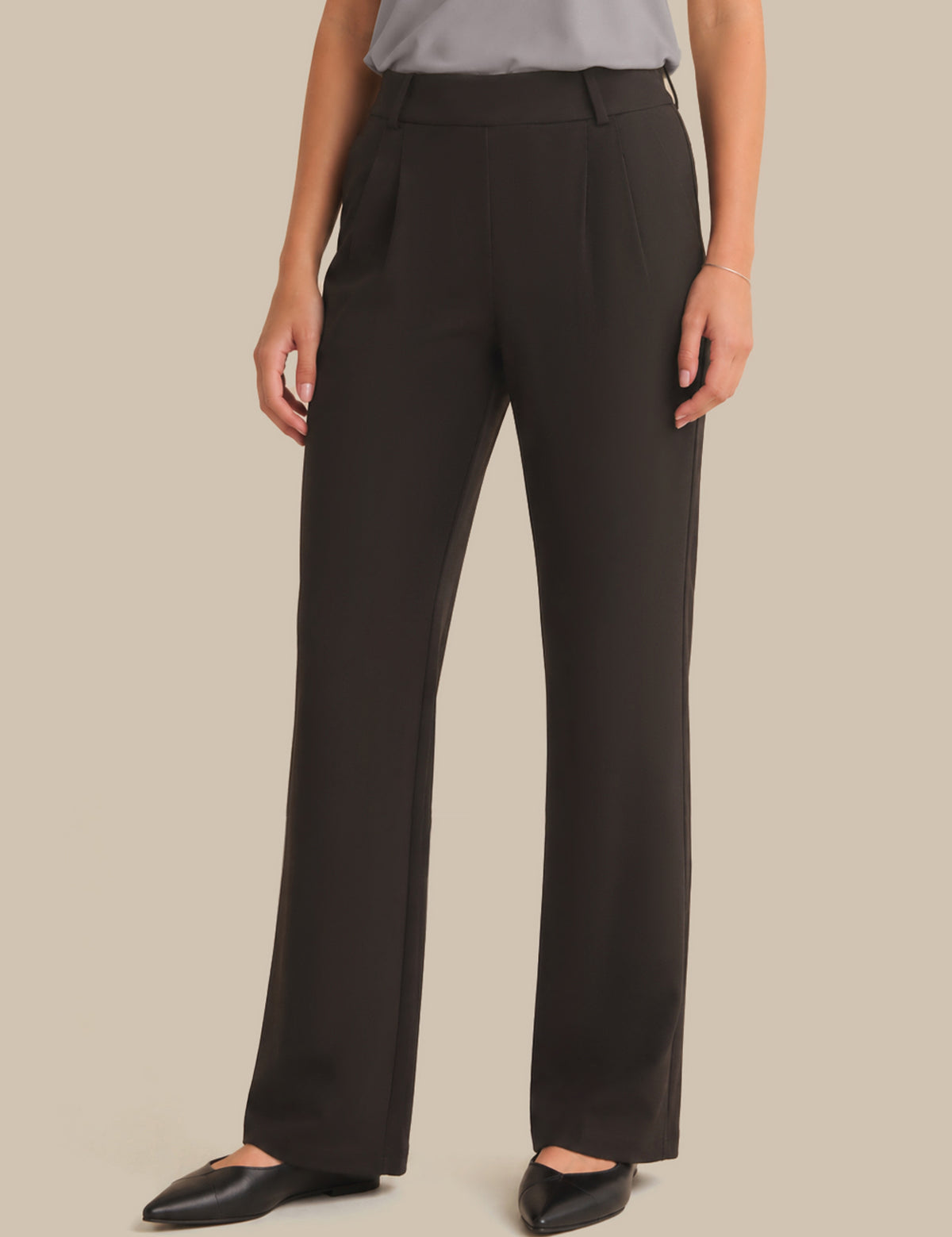A model wearing dark brown wide-leg trousers with a gray top and black pointed flats, shown from the waist down against a solid beige background.
