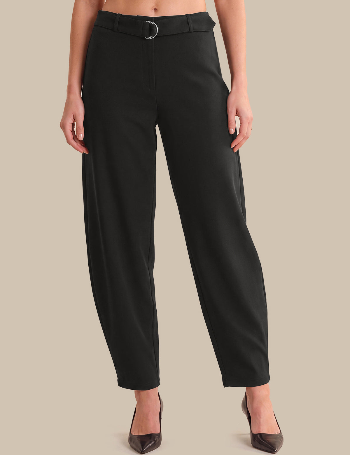 A model wearing black barrel-leg cropped trousers with a matching black belt featuring a silver-toned ring, paired with dark brown pointed-toe heels, set against a solid beige background.
