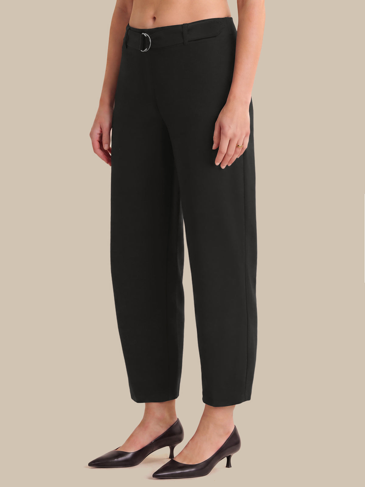 A model wearing black cropped trousers with a single silver-tone ring detail at the waistband, paired with black pointed-toe kitten heels, against a solid beige background.