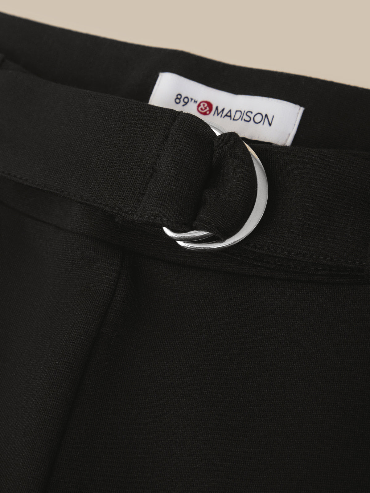 Close-up of a black skirt or dress waistband with a matching black belt and a large silver-tone metal O-ring, featuring a white interior label that reads 89th & MADISON.