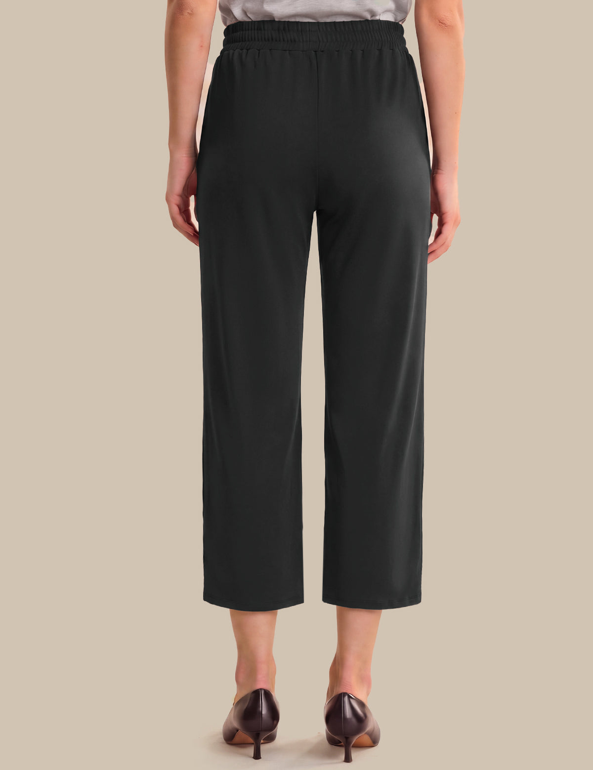 A model shown from the back wearing black cropped pants with a wide elastic waistband, paired with dark brown pointed-toe heels against a solid beige background.