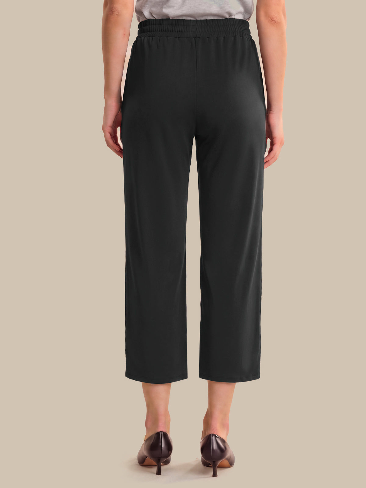 A model shown from the back wearing black cropped pants with a wide elastic waistband, paired with dark brown pointed-toe heels against a solid beige background.