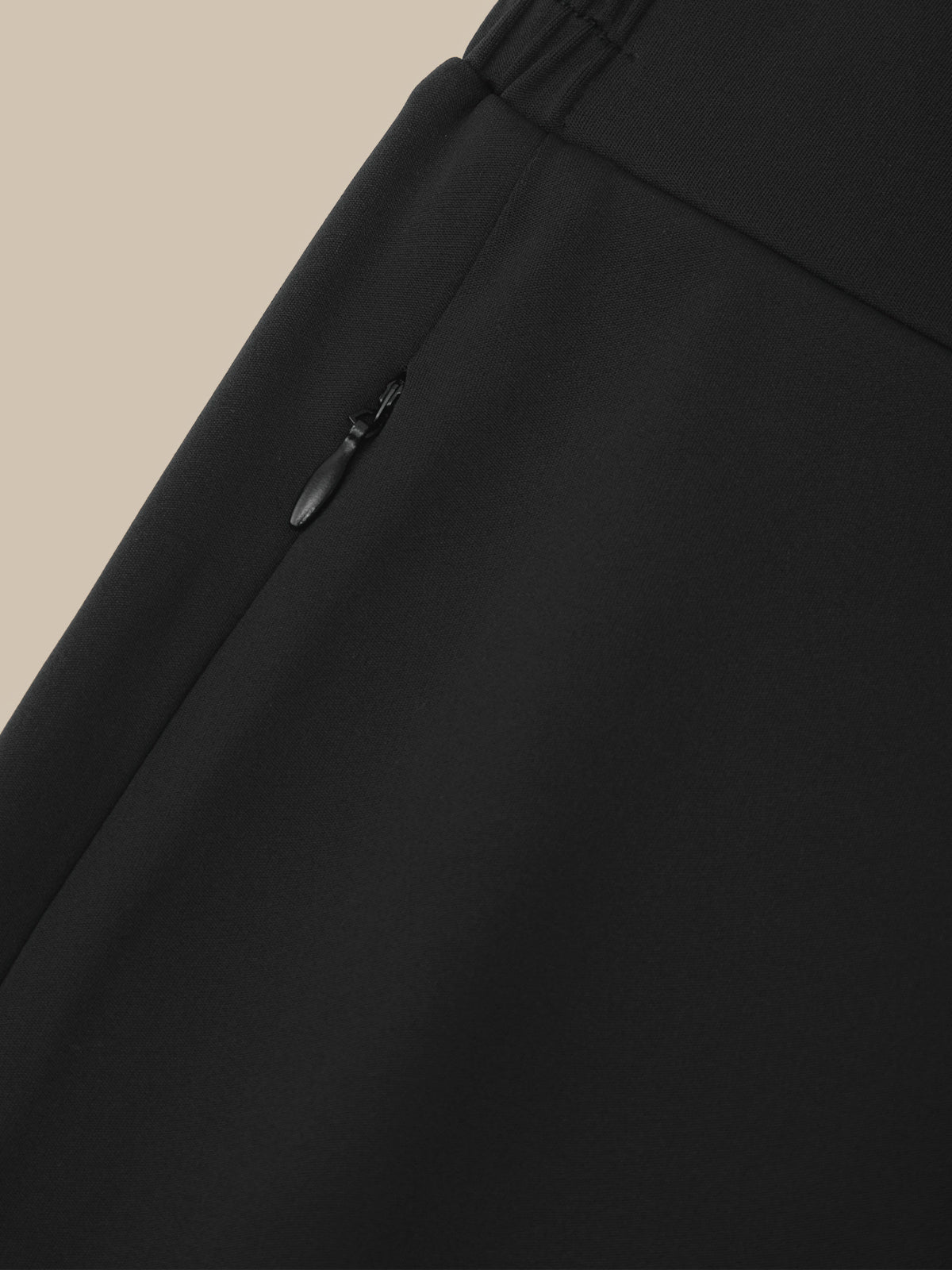 Close-up detail shot of black trousers showing an elastic waistband and a zippered pocket with a black zipper pull, set against a solid beige background.
