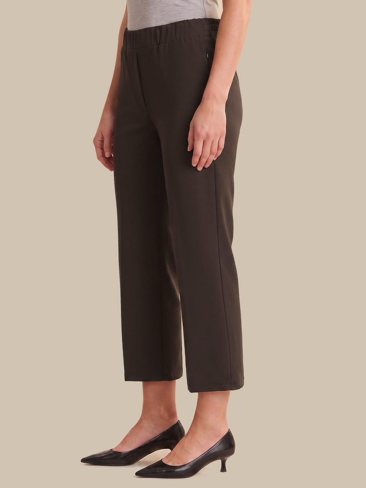 A model wearing dark brown cropped trousers with an elastic waistband, paired with black pointed-toe kitten heels, against a solid beige background.