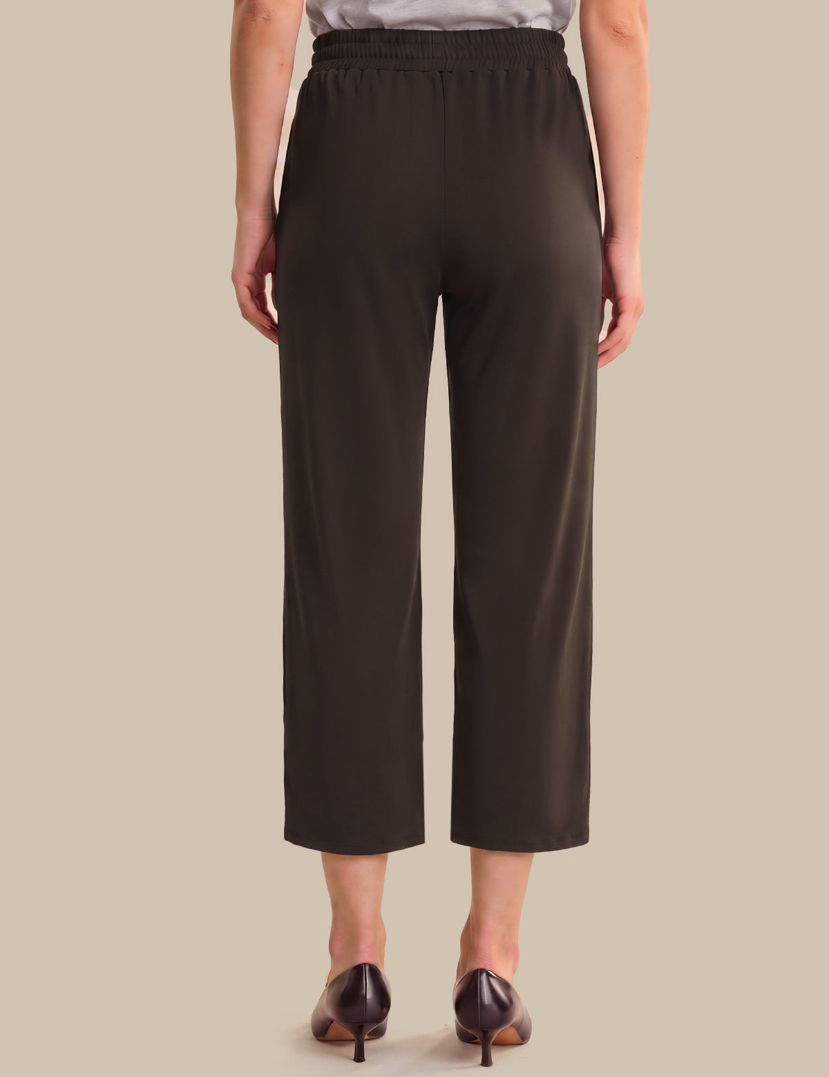 A model shown from the back wearing dark brown cropped trousers, a light gray top, and dark brown pointed-toe heels against a solid beige background.