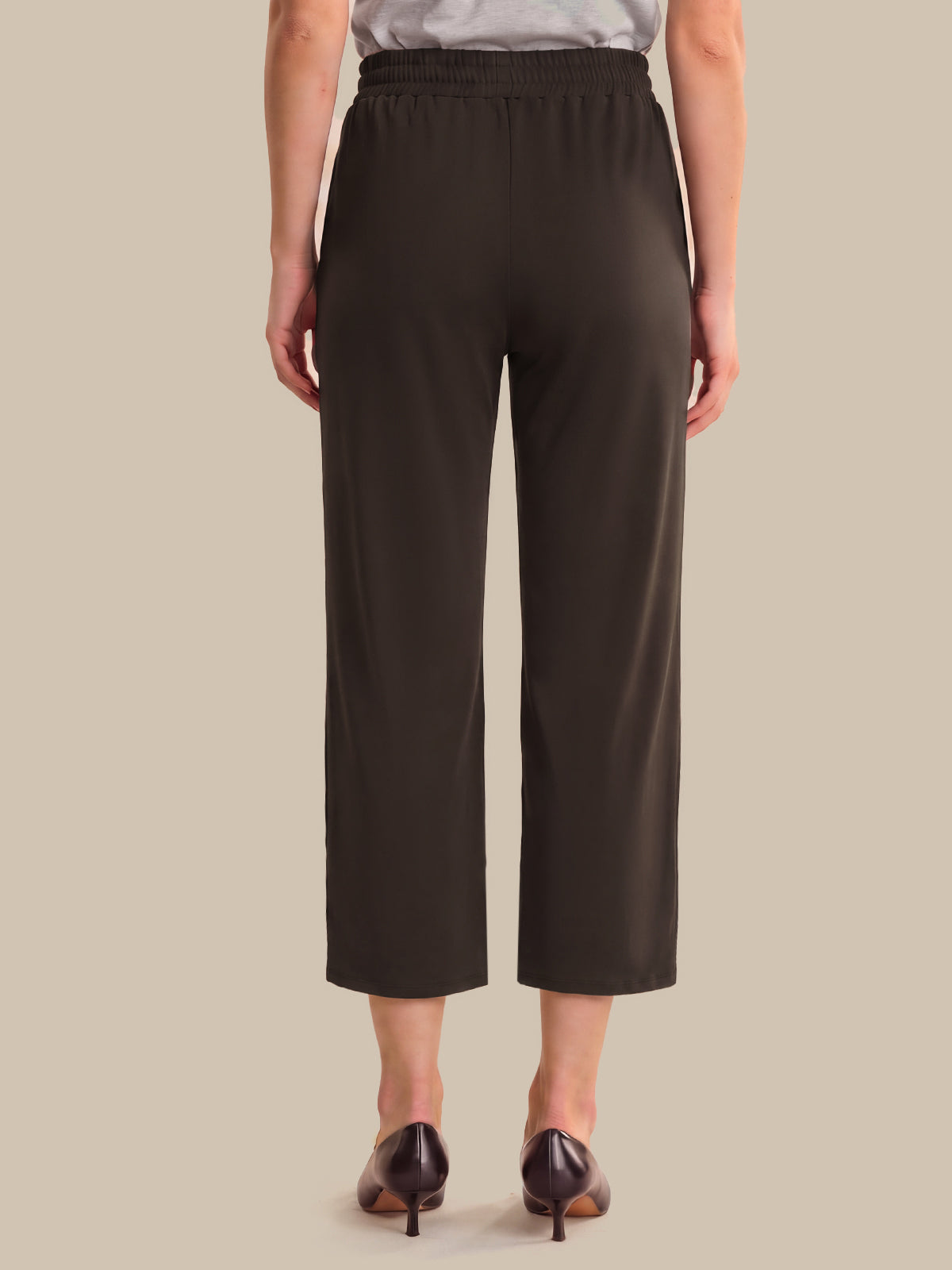 A model shown from the back wearing dark brown cropped trousers, a light gray top, and dark brown pointed-toe heels against a solid beige background.
