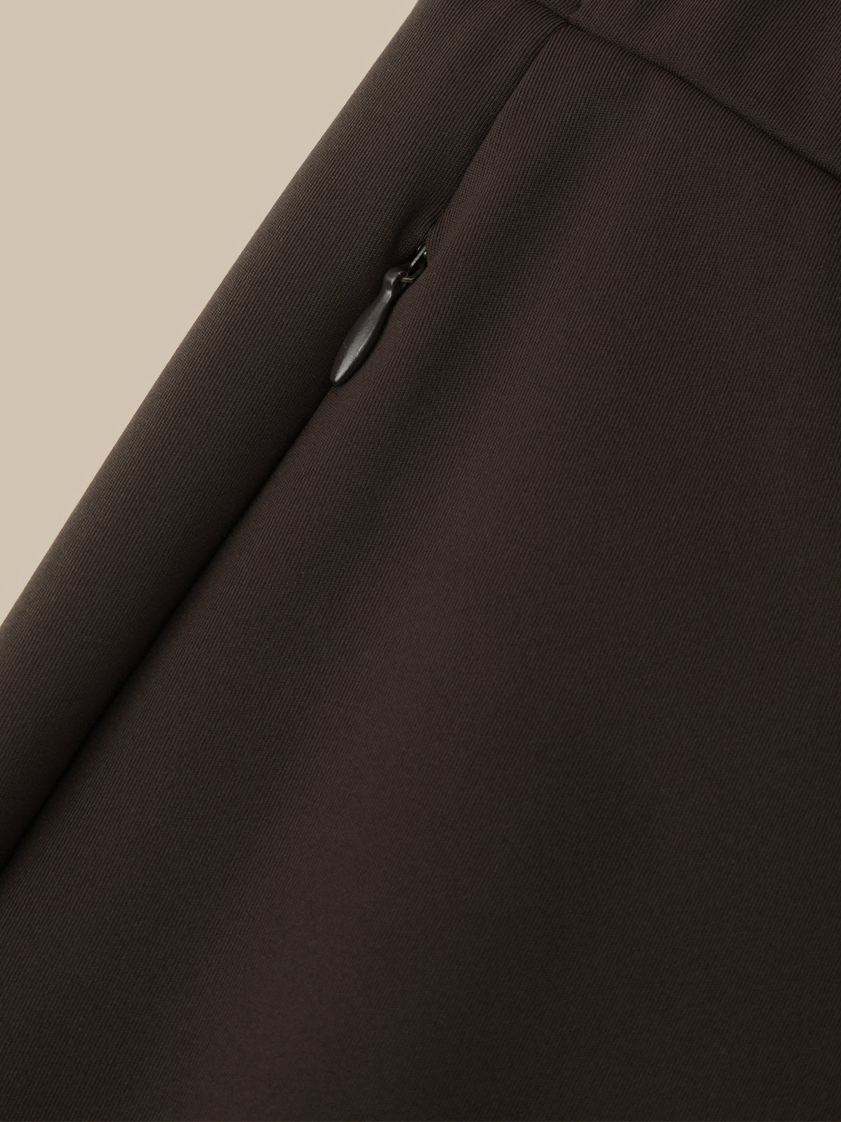 Close-up of a dark brown fabric garment showing a diagonal seam and a small black zipper pull on a light beige background.