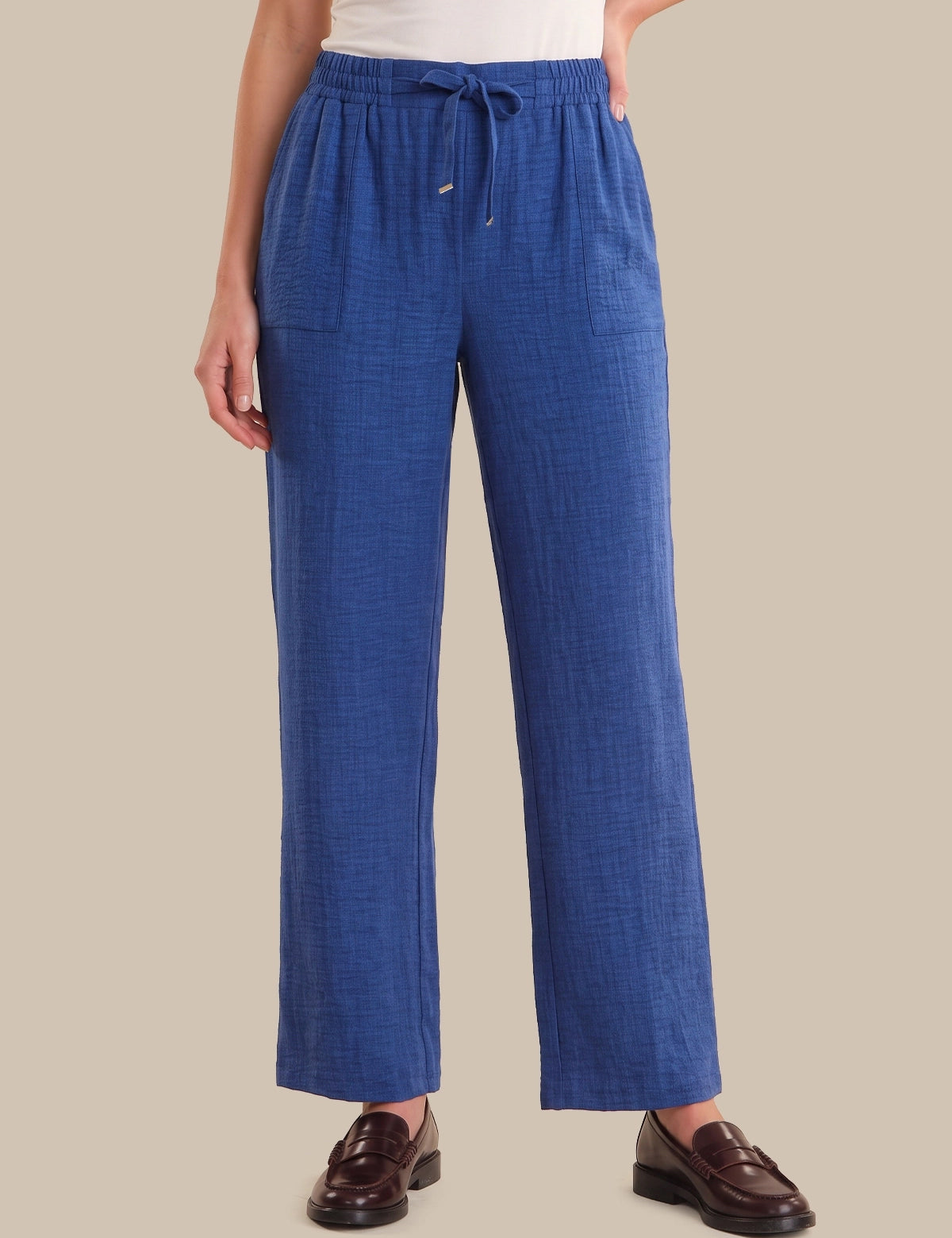 A person wearing blue wide-leg drawstring pants with a textured linen-like fabric, paired with a white top and brown leather loafers, against a solid beige background.
