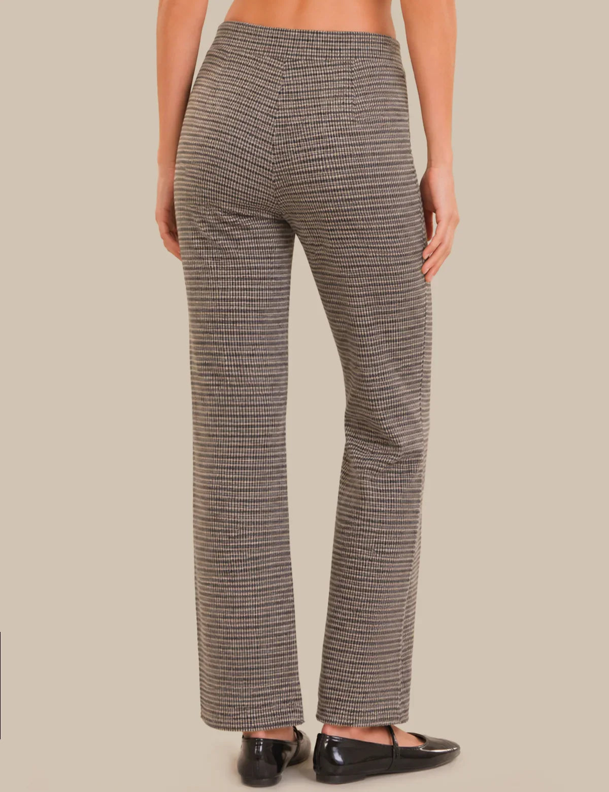 A person shown from the back wearing high-waisted, straight-leg trousers with a fine black and beige houndstooth pattern, paired with black flats, against a solid beige background.