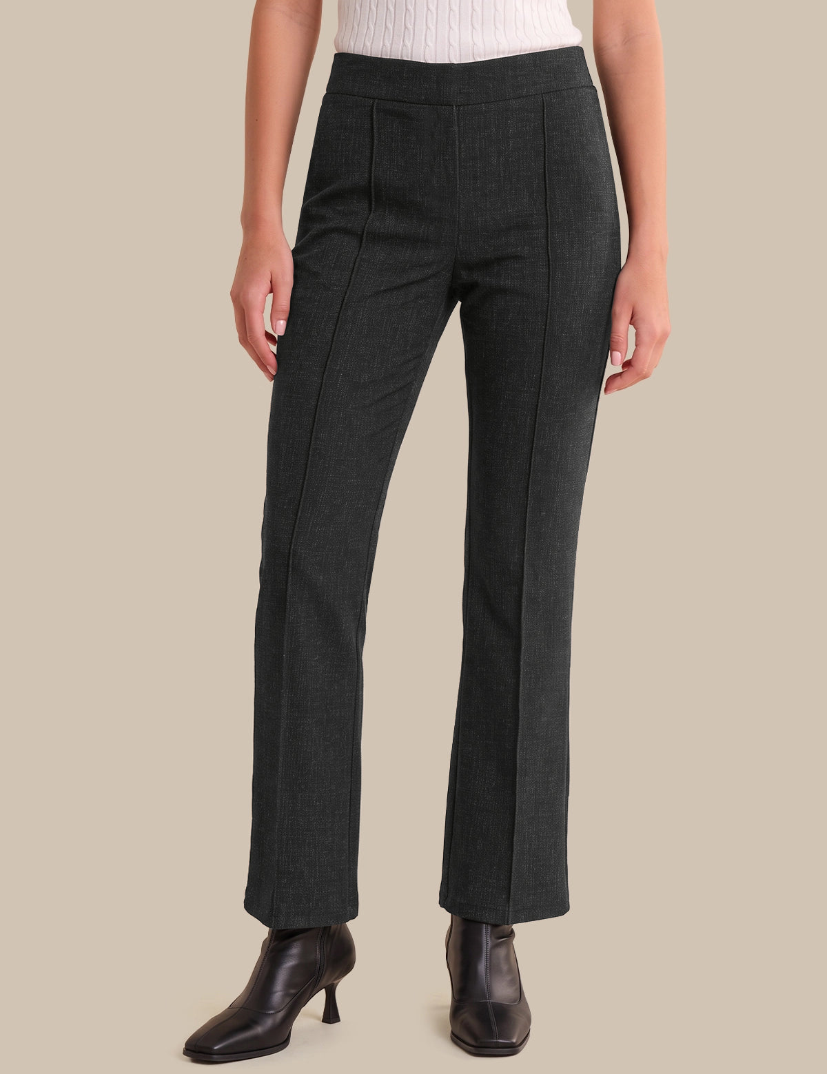 A model wearing dark charcoal gray straight-leg trousers, paired with a white ribbed top and black leather ankle boots, set against a solid beige background.