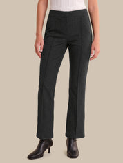 A model wearing dark charcoal gray straight-leg trousers, paired with a white ribbed top and black leather ankle boots, set against a solid beige background.
