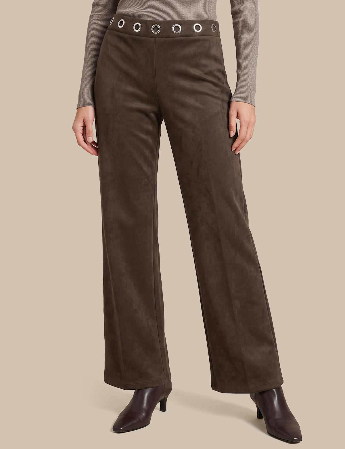 A model is wearing a pair of dark brown wide-leg trousers made of a suede-like material, featuring a matching waistband with silver-tone grommets. The model is also wearing a taupe long-sleeve top and dark brown heeled ankle boots, set against a solid beige background.