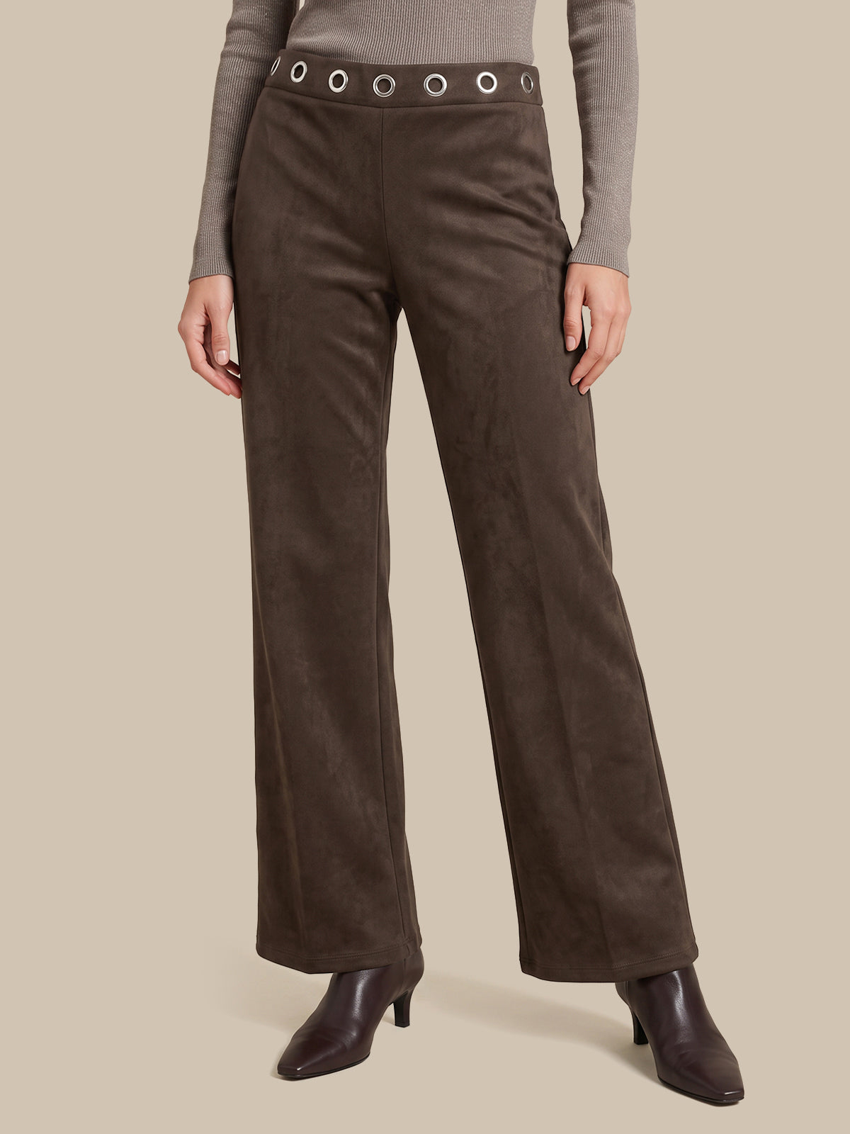A model is wearing a pair of dark brown wide-leg trousers made of a suede-like material, featuring a matching waistband with silver-tone grommets. The model is also wearing a taupe long-sleeve top and dark brown heeled ankle boots, set against a solid beige background.