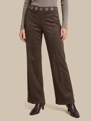 A model is wearing a pair of dark brown wide-leg trousers made of a suede-like material, featuring a matching waistband with silver-tone grommets. The model is also wearing a taupe long-sleeve top and dark brown heeled ankle boots, set against a solid beige background.