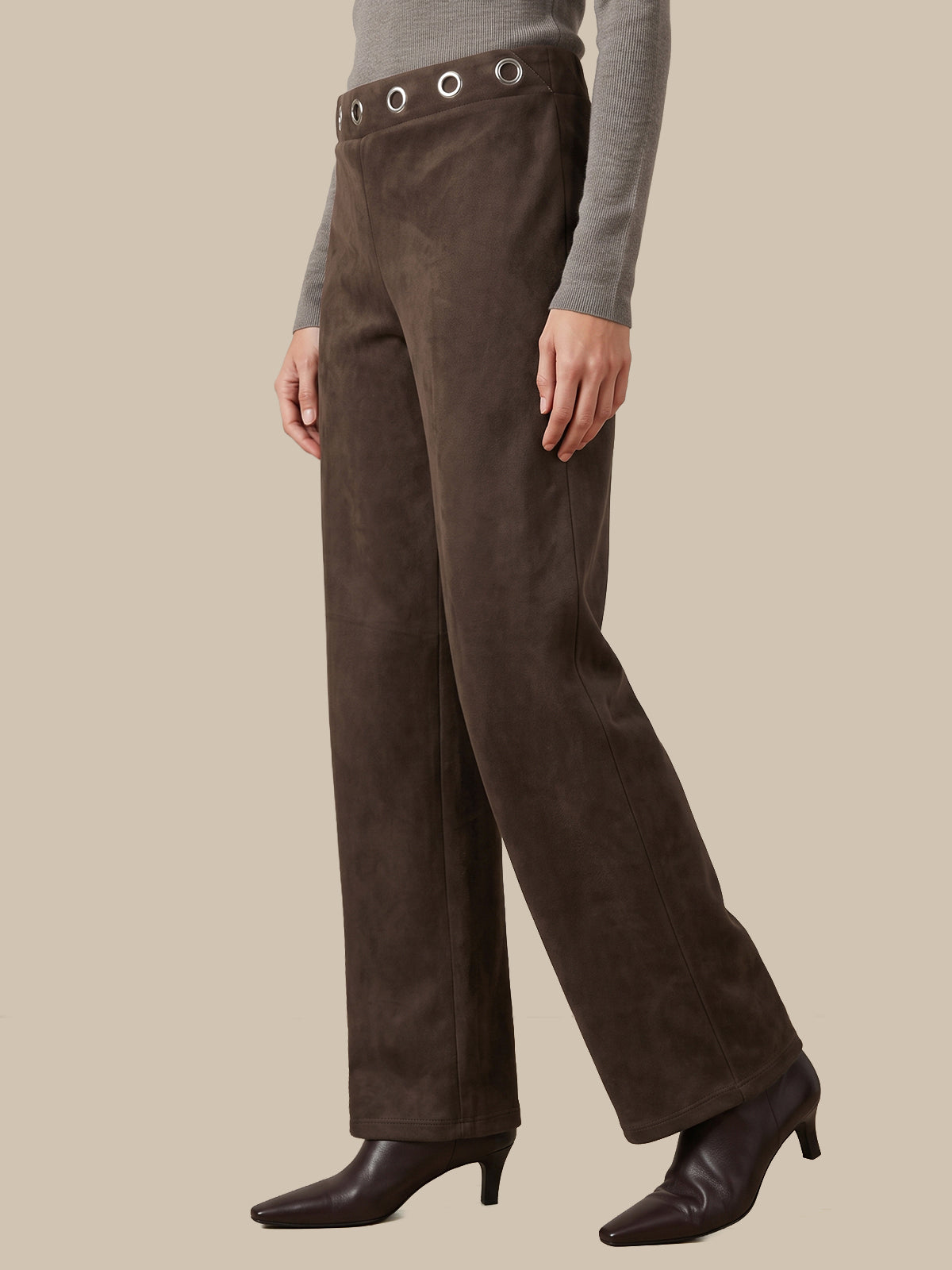 A model wearing dark brown wide-leg trousers with a matching waistband belt and silver-tone grommets, styled with a gray long-sleeve top and dark brown pointed-toe heeled boots against a solid beige background.