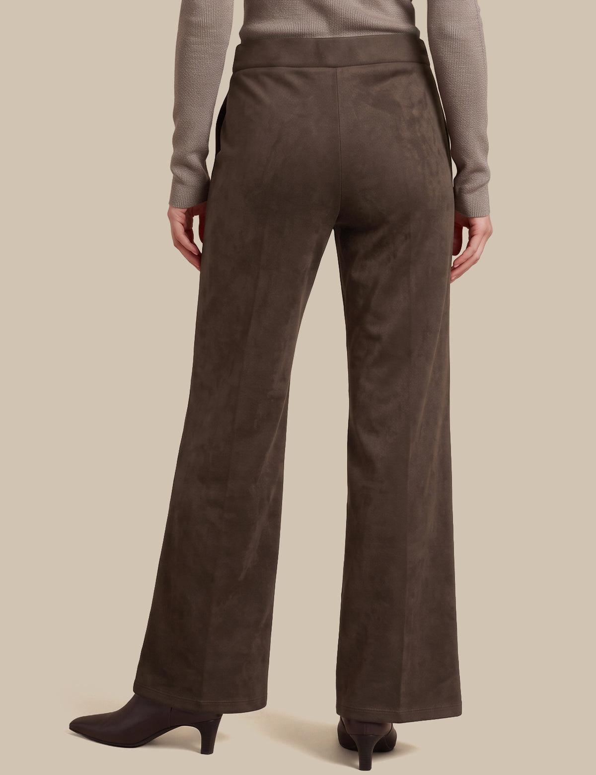 A person shown from the back wearing dark brown wide-leg trousers, a taupe long-sleeve top, and dark heeled boots against a plain beige studio background.