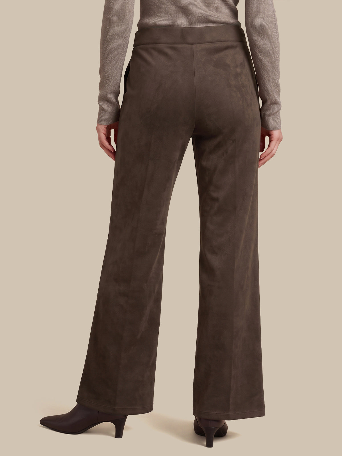 A person shown from the back wearing dark brown wide-leg trousers, a taupe long-sleeve top, and dark heeled boots against a plain beige studio background.