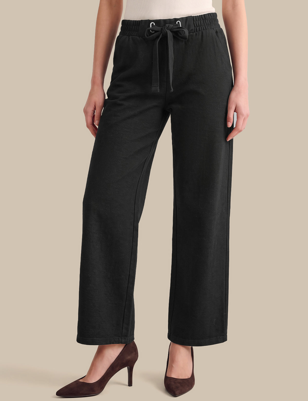 A model wearing black wide-leg cropped pants with an elastic waistband and drawstring, paired with dark brown pointed-toe heels against a solid beige background.