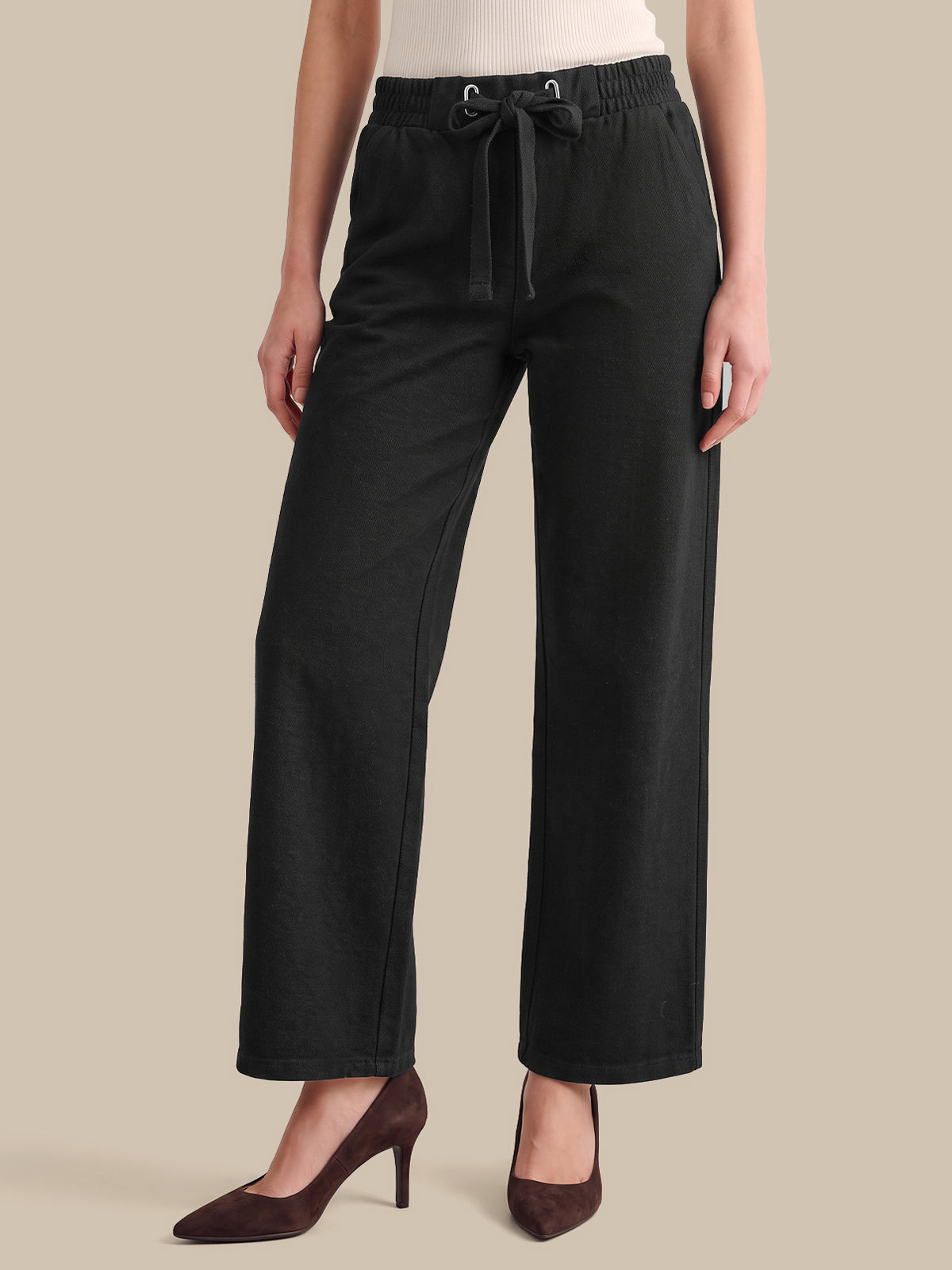 A model wearing black wide-leg cropped pants with an elastic waistband and drawstring, paired with dark brown pointed-toe heels against a solid beige background.