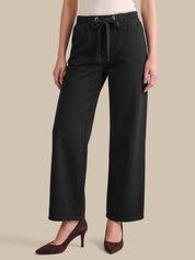 A model wearing black wide-leg cropped pants with an elastic waistband and drawstring, paired with dark brown pointed-toe heels against a solid beige background.
