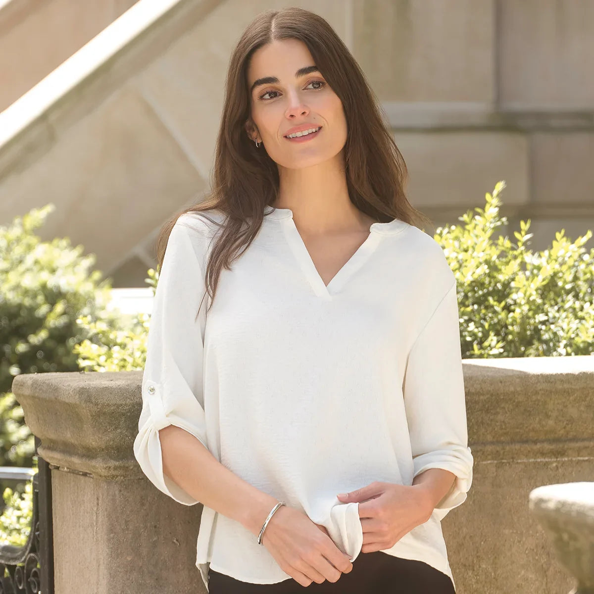 A woman wearing a white long-sleeve blouse with a split V-neckline, standing outdoors in front of a stone wall and greenery.